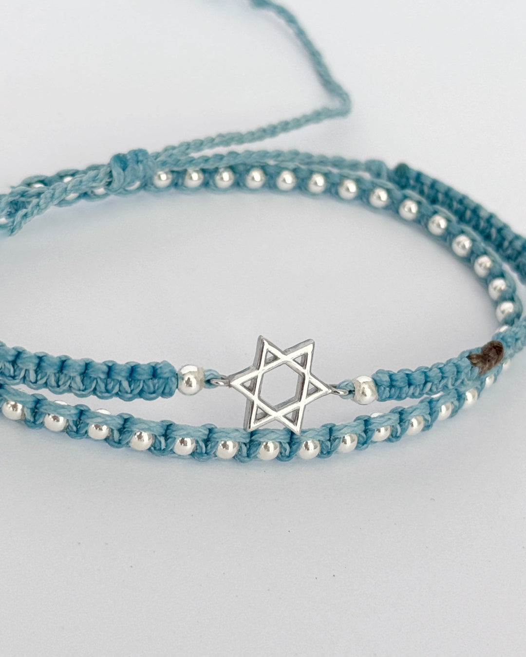 צמיד Shield הוא צמיד מקרמה כפול בגוון תכלת-שמיים הכולל תליון מגן דוד מינימליסטי וחרוזי כסף 925. העיצוב משלב טקסטורה רכה של החוט עם אלמנטים מכסף, ויוצר שקט צורני שיש בו גם הגנה וגם נוכחות.