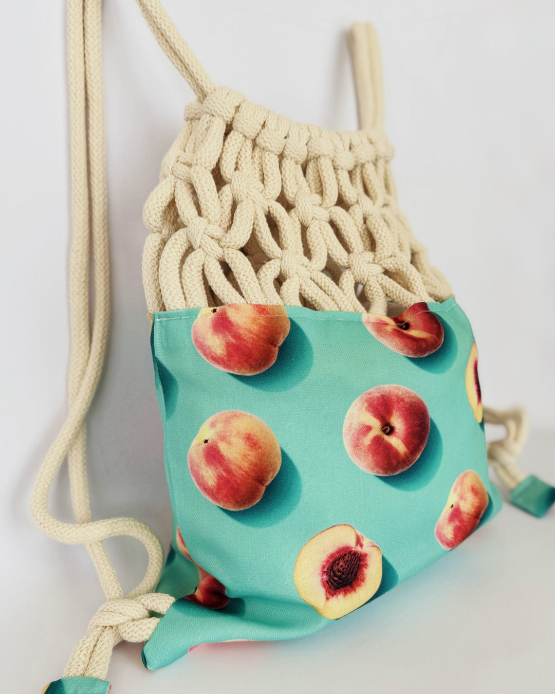 Handmade macramé backpack with cream macramé top and turquoise bottom featuring a peach fruit print – Tropica Nero design, tropical and light in style.
